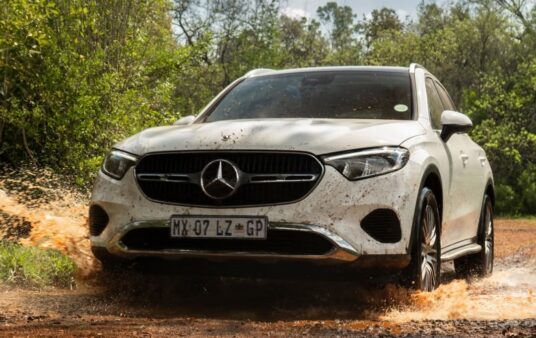 Action shot of a white Mercedes-Benz GLC Coupe splashing aggressively through a deep mud puddle, demonstrating its impressive 4x4 performance on dirt tracks