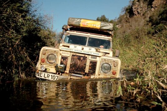A classic Land Rover Defender navigating a deep river crossing in the bush, a challenging obstacle that can easily ruin an engine if recovery gear and snorkels are not properly fitted.